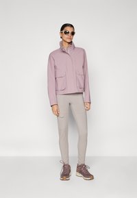 Lilac waterproof jacket with a high collar, two front pockets, paired with light grey fitted pants and dark brown hiking shoes.