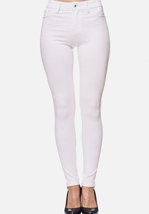 Hvide skinny jeans med høj talje båret af en stående kvinde, parret med sorte højhælede sko, på en ensfarvet hvid baggrund.