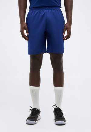 Mann trägt königsblaue Sportshorts, weiße Sportsocken und schwarze Laufschuhe und steht vor einem schlichten Hintergrund.