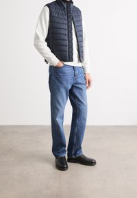 Granatowa, ocieplana kamizelka na lekkim szarym swetrze, w zestawieniu z niebieskimi dżinsami i czarnymi butami. Kamizelka ma wysoki kołnierz i zamek błyskawiczny.