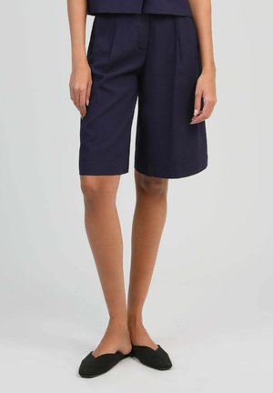 Femme debout portant un short tailleur bleu marine mi-cuisse et des chaussures plates noires à bout pointu, slip-on, sur un fond gris clair.