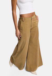 Pantaloni a gamba larga color sabbia realizzati in tessuto liscio, con vita alta, zip frontale e tasche laterali. Abbinati a scarpe beige a punta.