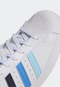 White sneaker with a textured toe cap, featuring blue and light blue zigzag striped accents. Laces are white, with perforations on the sides.