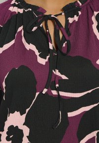 Blusa viola e nera con un motivo floreale, caratterizzata da un colletto arricciato e un dettaglio con fiocco sul davanti per la regolazione. Tessuto texturizzato.