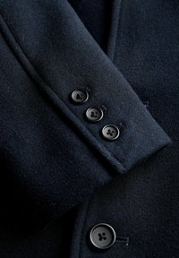 Temno mornarsko volneno plašč z rokavom in sprednjo ploščo, na kateri so vidni štirje črni gumbi, prikazuje natančne šive in teksturirano tkanino.