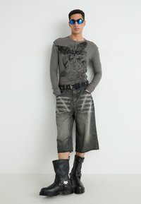 Graues Langarmshirt mit Grafikdesign, dunkle Jeansshorts mit verblasstem Muster, schwarze Lederstiefel mit Schnallen und strukturierten Sohlen.