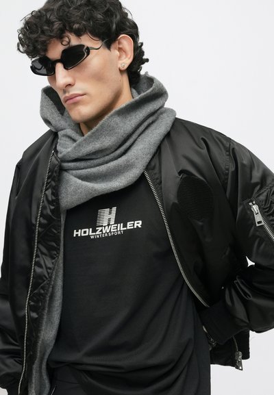 Μαύρο bomber jacket με φερμουάρ, γκρι πλεκτό κασκόλ, μαύρο T-shirt με στάμπα "HOLZWEILER WINTERSPORT" και κομψά μαύρα γυαλιά ηλίου.