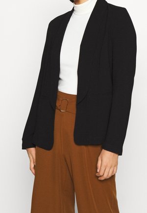 Person trägt einen schwarzen Blazer, ein weißes Rollkragentop und hoch taillierte braune Hose mit Gürtelschnalle vor einem schlichten weißen Hintergrund.
