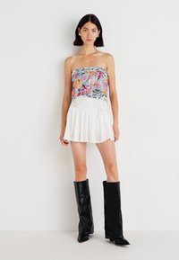 Haut sans bretelles avec motif floral coloré et franges, associé à une jupe blanche évasée et des bottes en cuir noir jusqu'aux genoux.