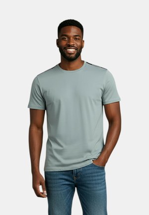 Uomo sorridente con indosso una maglietta a maniche corte grigio chiaro e jeans blu, in piedi con una mano in tasca contro uno sfondo semplice.