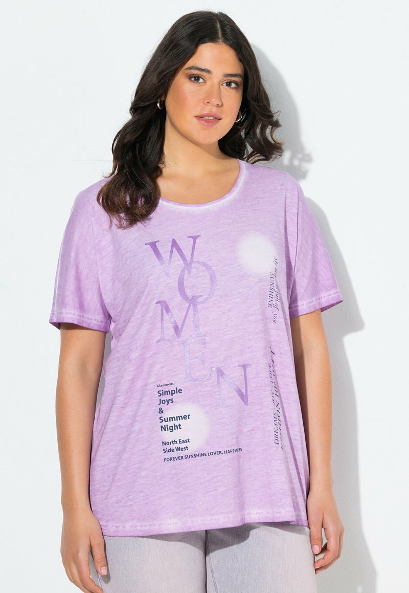 Mujer vistiendo una camiseta lavanda con "MUJERES" y palabras motivacionales impresas en diferentes tipografías, combinada con pantalones de colores claros.