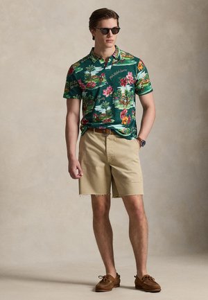 Jeune homme portant un polo vert à motifs floraux, un short beige, des chaussures bateau marron, des lunettes de soleil et une montre-bracelet, debout avec une main dans la poche.