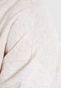 Sudadera de color beige claro con una textura de tela que presenta un sutil patrón jaspeado, destacando un diseño de costura del hombro redondeado.