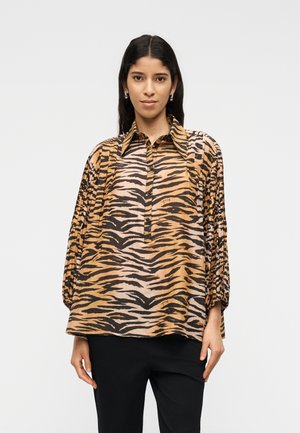 Hosszú, sötét hajú nő laza, tigriscsíkos blúzt visel pliszírozott ujjal és fekete nadrágot, egy egyszínű háttér előtt áll.