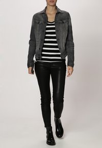 Frau, die eine graue Jeansjacke, ein schwarz-weiß gestreiftes Oberteil, schwarze Lederhosen und schwarze Ankle Boots trägt und eine kleine schwarze Clutch hält.