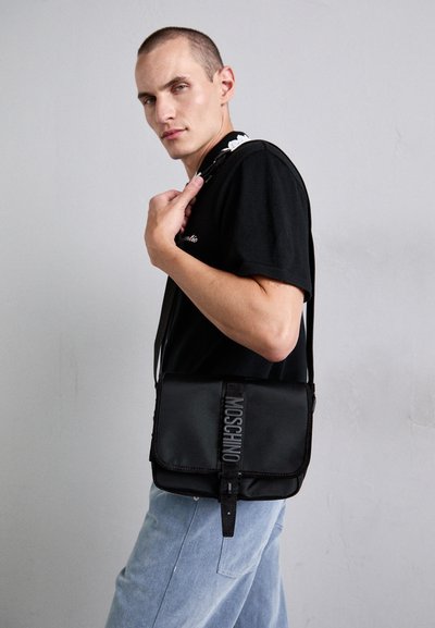 Băiat tânăr îmbrăcat cu o cămașă neagră și blugi de culoare bleumarin deschis, purtând o geantă de umăr neagră Moschino, în fața unei pereți gri uni.