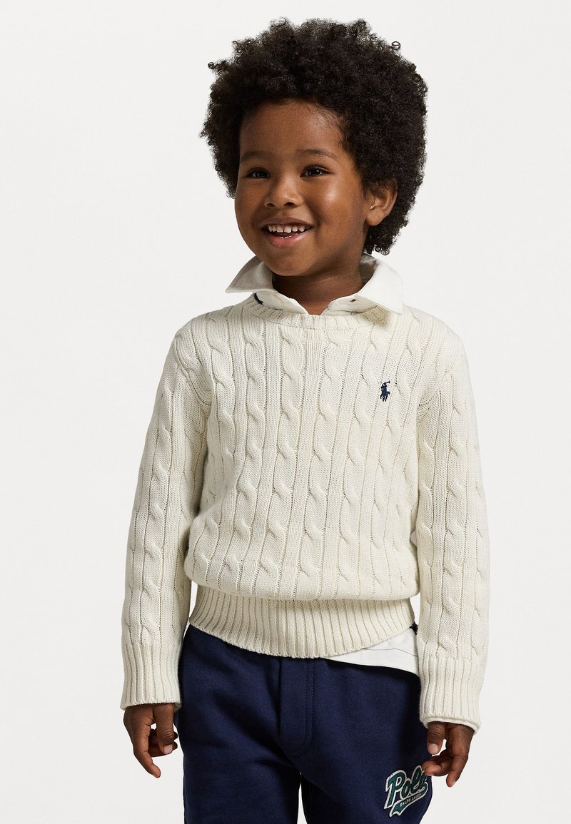 Krämfärgad kabelstickad tröja med ribbad nederkant och manschetter, lager på lager över en vit skjorta med krage. Bärs med marinblå byxor med en logotyp.