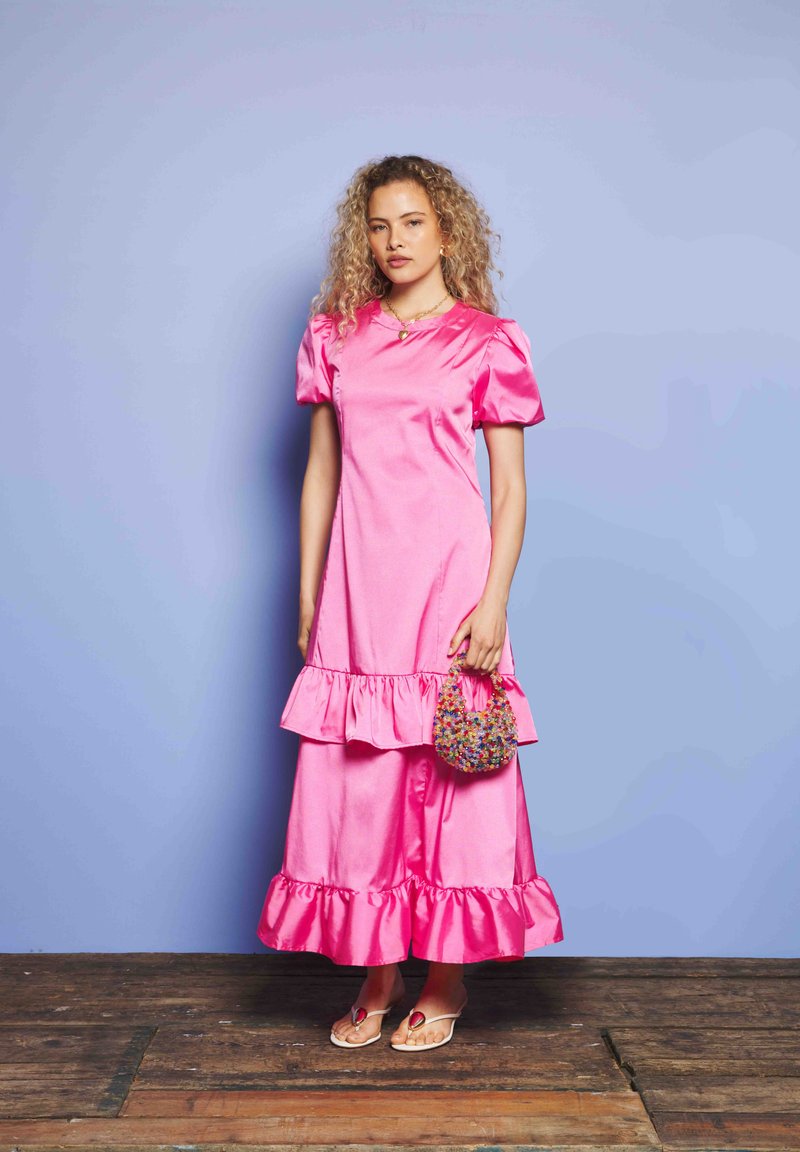 Ženska s kodrastimi lasmi, oblečena v svetlo roza obleko z volančki, drži barvno perličasto torbico in stoji na lesenem tleh pred modro steno.