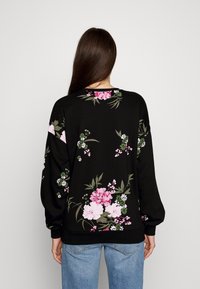Mujer con cabello largo castaño que lleva una sudadera negra con patrones florales en rosa y blanco, combinada con jeans de color azul claro, de espaldas.