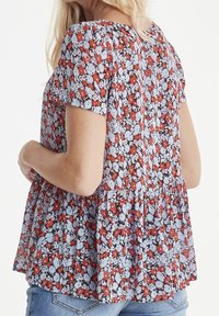 Femme portant un blouse florale ample à manches courtes avec des fleurs rouges et bleu clair, associée à un jeans bleu, vue de derrière.