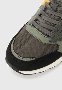 Gros plan sur l'avant de la sneaker avec un maillage vert olive, des superpositions en daim, des lacets gris et une semelle en caoutchouc noire avec une bande médiane blanche.