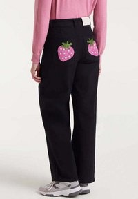 Persona que lleva pantalones negros con diseños de fresas rosas en los bolsillos traseros, combinados con una blusa rosa de manga larga y zapatillas grises.