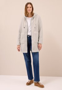 Manteau gris et duveteux avec une capuche et deux poches avant. Fermeture éclair et cordons ajoutent une touche au design. Porté par-dessus une chemise blanche et un jean bleu.