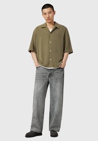 Korte mouwen olijfgroene overhemd met knopen, relaxte pasvorm; gecombineerd met losse grijze jeans en donkere schoenen. Subtiele merklogo op het shirt.