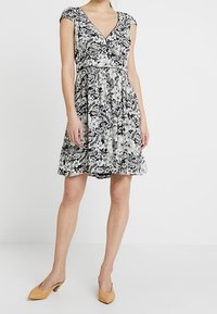 Žena v černobílé květinové krátkosrsté šaty s výstřihem do V, dosahující ke kolenům, se světlými nízkými jehlovými botami, stojící na jednotném pozadí.