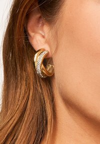 Orecchini a cerchio in oro con un design curvo, caratterizzati da sezioni lisce e lucide, e adornati con una fila di piccoli cristalli trasparenti.