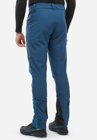 Homme portant un pantalon de plein air bleu avec des détails de fermeture éclair sur le côté et des chaussures de randonnée noires, se tenant devant un fond blanc uni.
