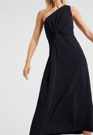 Nainen yllään virtaava musta yhden olkapään mekko, jossa on kokoontaitettu vyötärö, kuvattuna kaulasta nilkkoihin yksinkertaisella taustalla.