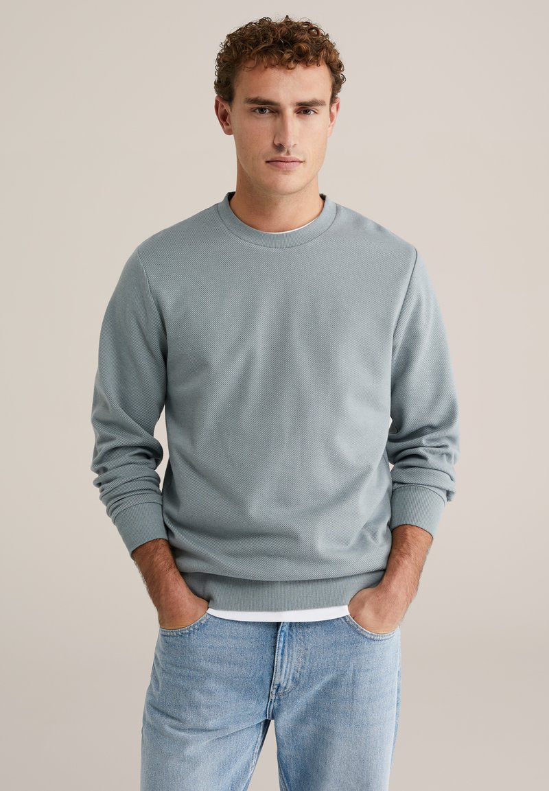 Hele sinine ümarselja dressipluus soonikkoes mansettide ja äärega, millel on tekstuurse kangaga disain. Kandmisel valge särgi kohal ja heledate teksadega.