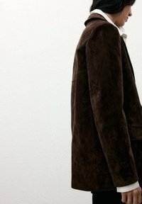 Blazer in suede marrone con una vestibilità rilassata, caratterizzato da un colletto a lancia e spalle strutturate. Accompagnato da una camicia bianca sotto.