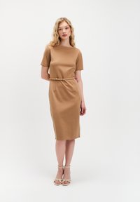 Młoda kobieta z falującymi blond włosami, ubrana w dopasowaną, beżową sukienkę do kolan z krótkim rękawem, cienkim paskiem i beżowymi sandałami na obcasie z odkrytymi palcami.