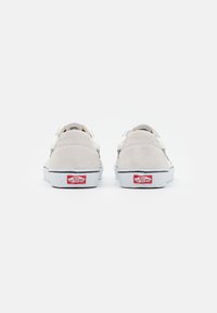 Szare zamszowe sneakersy w niskim kroju z białymi skórzanymi akcentami, białą gumową podeszwą i czerwonym logo na pięcie. Widok z tyłu, minimalistyczny design.