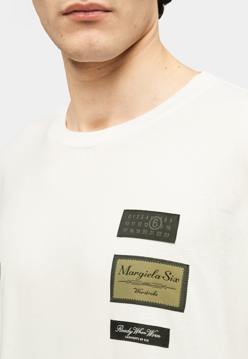 T-shirt en coton blanc avec trois étiquettes en tissu vert foncé et noir, comportant des motifs textuels et numériques, positionnées sur la poitrine.