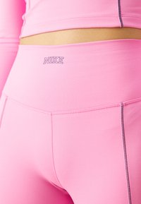 Des leggings de sport roses présentent une taille haute avec un logo subtil, une texture lisse et des coutures contrastantes le long des seams.
