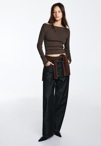 Top marrone con maniche lunghe e scollatura a barca abbinato a jeans in denim scuro a vita alta. L'outfit include un maglione marrone annodato in vita.