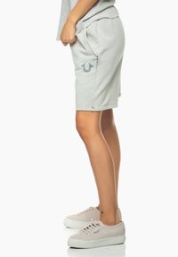 Světle šedé šortky vyrobené z měkké tkaniny, vybavené bočními kapsami a logem na levé noze. V kombinaci se světlými teniskami.