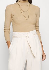 Pull à col roulé côtelé beige associé à un pantalon taille haute crème avec une ceinture en tissu et des jambes larges, créant un look texturé et superposé.