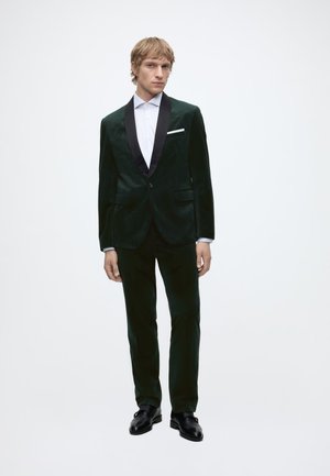 Man wearing dark green velvet tuxedo with black satin lapels, white dress shirt, white pocket square, and black dress shoes on a plain background.