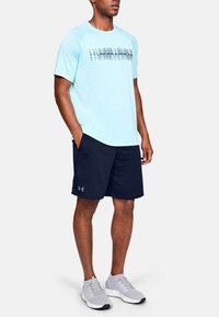 Ljusblå atletisk T-shirt med "Under Armour"-logotyp, i kombination med marinblå shorts och grå löparskor. Slätt tyg, avslappnad design.