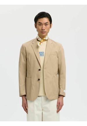 Uomo che indossa un blazer beige sopra una t-shirt grafica color crema e pantaloni bianchi larghi, con sciarpa gialla a motivi annodata intorno al collo su sfondo bianco.