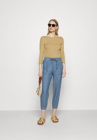 T-shirt bege ajustado de manga longa, calças azuis com cordão e bolsos, sandálias amarelas e óculos de sol pretos. De pé contra um fundo branco.
