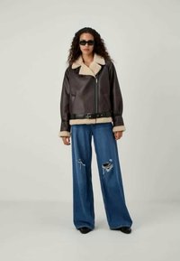 Ruda odinė striukė su puriu smėlio spalvos apykakle ir rankogaliais, užtrauktukas priekyje ir diržo juosmeniui; dėvima kartu su mėlynomis plačiomis džinsinėmis kelnėmis su nusidėvėjusiomis detalėmis.