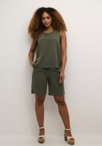 Olivenfarvet to-delt outfit med en ærmeløs top og knælange shorts, lavet af et tekstureret stof. Fodtøjet er lysefarvet med en tyk hæl.