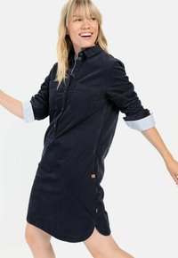 Koszula sukienkowa w kolorze granatowym, uszyta z miękkiego kordurowego materiału, z dwiema przednimi kieszeniami, rękawami z paskami z mankietami oraz zaokrąglonym dołem z bocznymi rozcięciami.