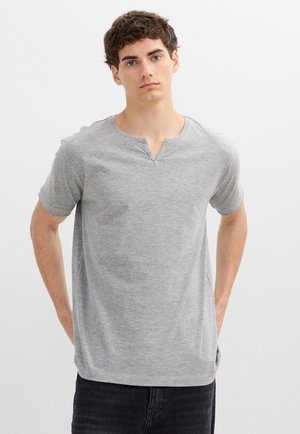 Joven con cabello castaño corto vistiendo una camiseta Henley gris de manga corta y pantalones negros, de pie contra un fondo blanco liso.