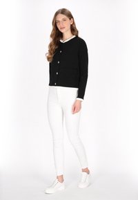 Cardigan negru scurt cu guler rotund, buzunar frontal și închidere cu nasturi din alamă, purtat peste un top alb și asortat cu pantaloni albi.